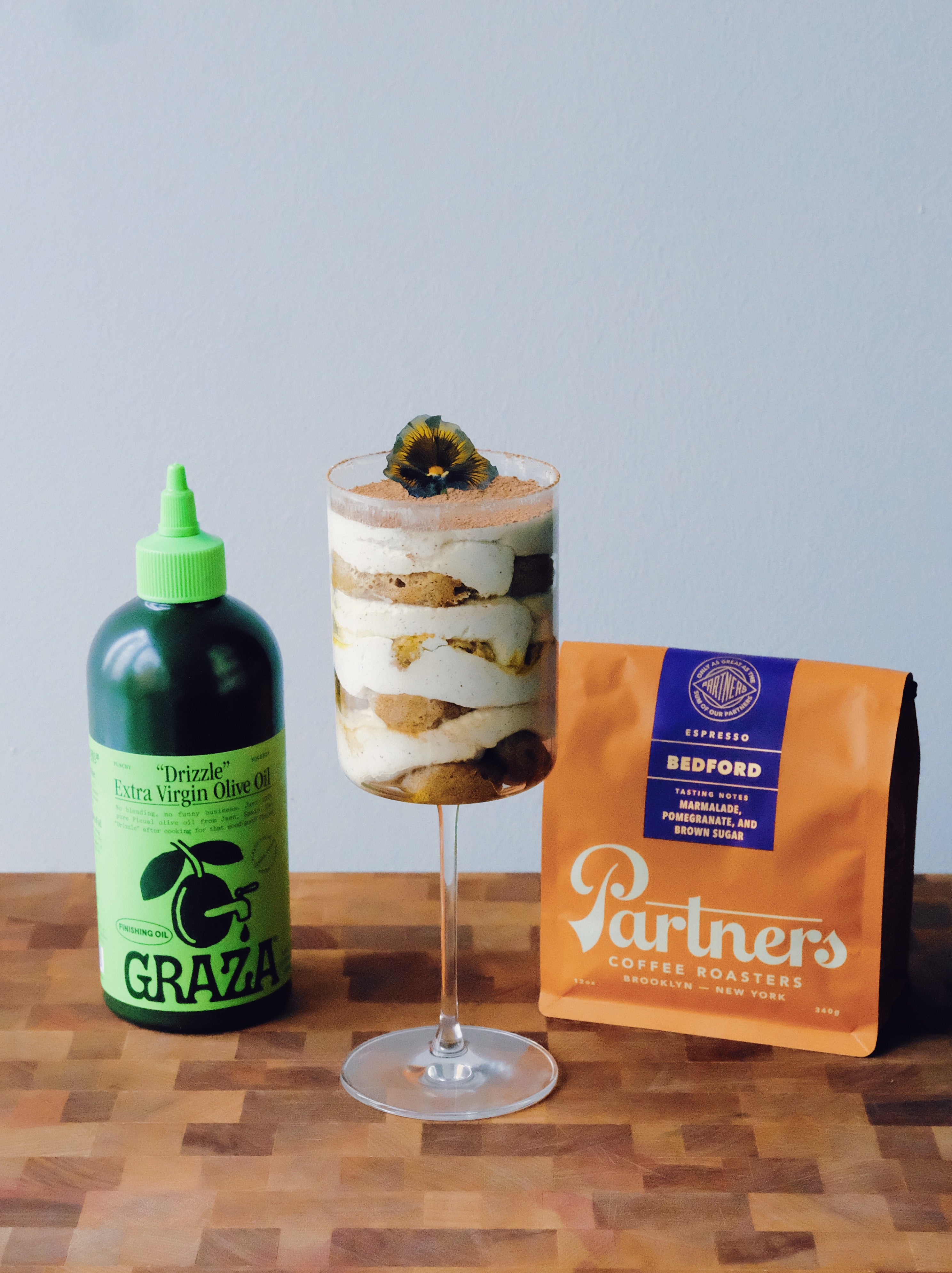 A bottle of green Graza olive oil sits to the left. In the middle there is a wine glass with layered Tiramisu topped with an edible flower. On the right an orange bag of Partners Coffee sits, labeled "Bedford"