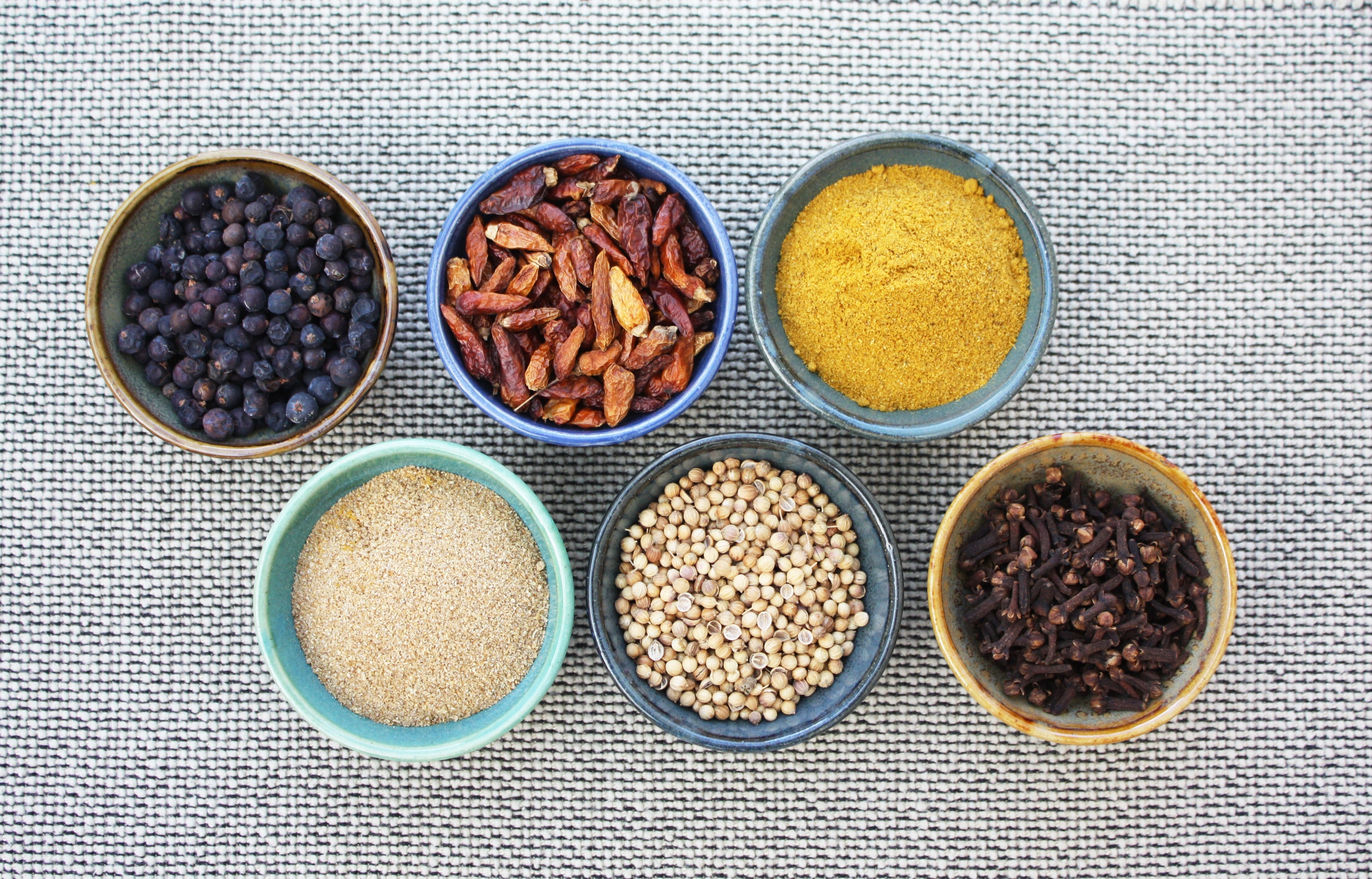 Top-down view of six small ceramic bowls holding various whole and ground spices and dried chilies on woven fabric