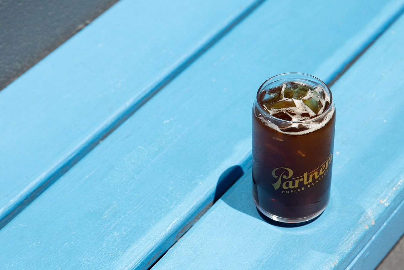 Glass of cold brew on a bright blue wooden bench, top-down view showing ice and a Partners logo on the glass