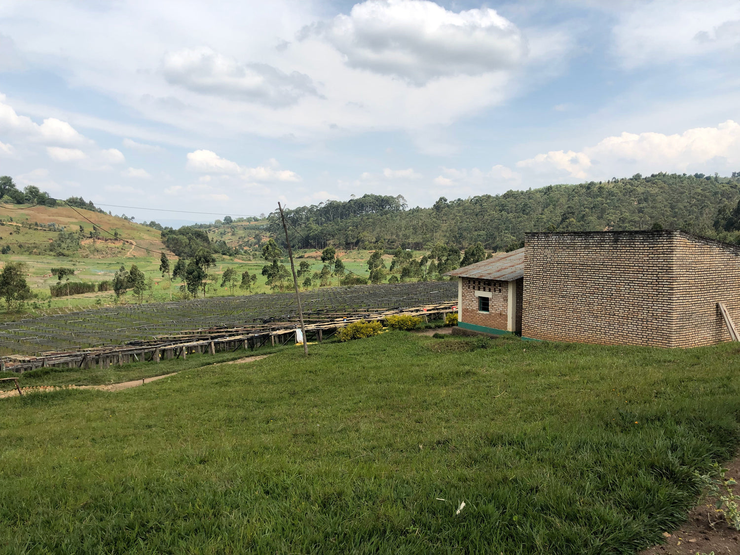Coffee drying beds and a brick processing building on a grassy hillside at Gahahe farm in Burundi under a partly cloudy sky