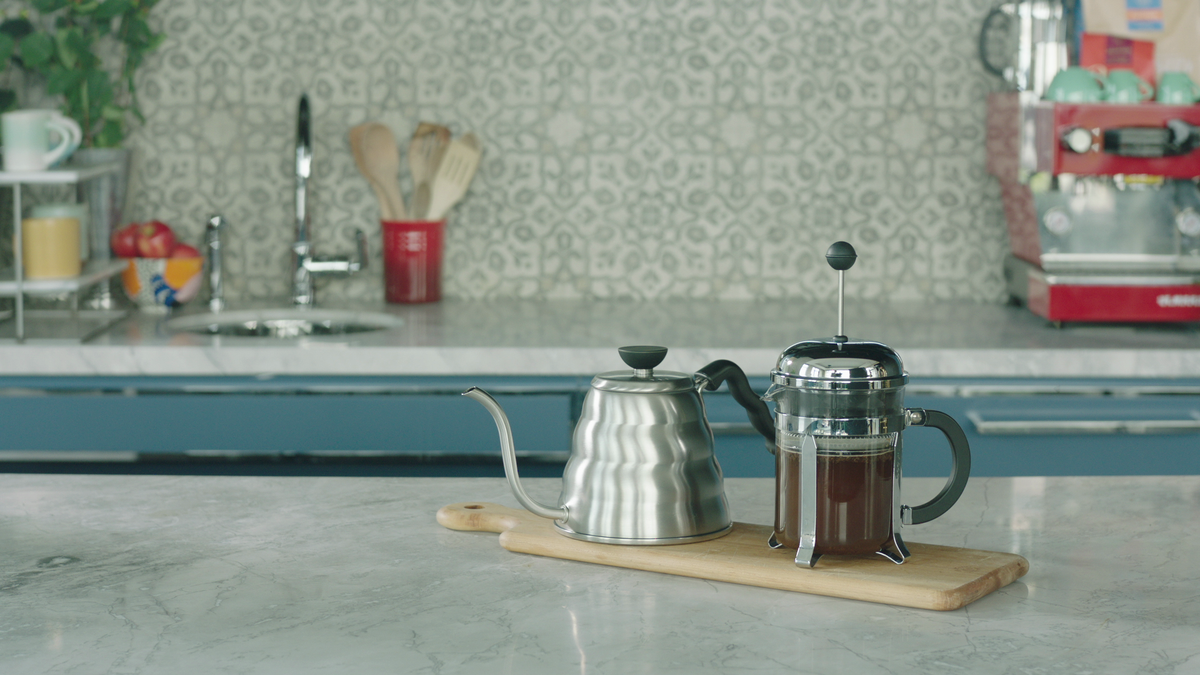 French press brewing coffee beside a stainless steel gooseneck kettle on a cutting board on a marble countertop in a bright kitchen