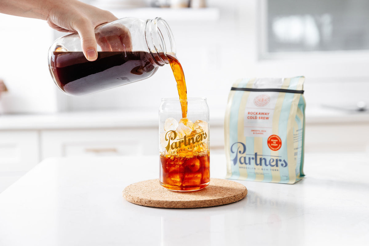 Jar pouring Rockaway Cold Brew into an ice filled Partners glass beside a Rockaway Cold Brew bag on a white counter