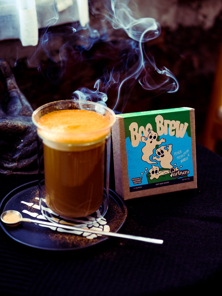 Steaming Spooky Butter Latte beside a Boo Brew instant coffee box on a dark table with a saucer and spoon