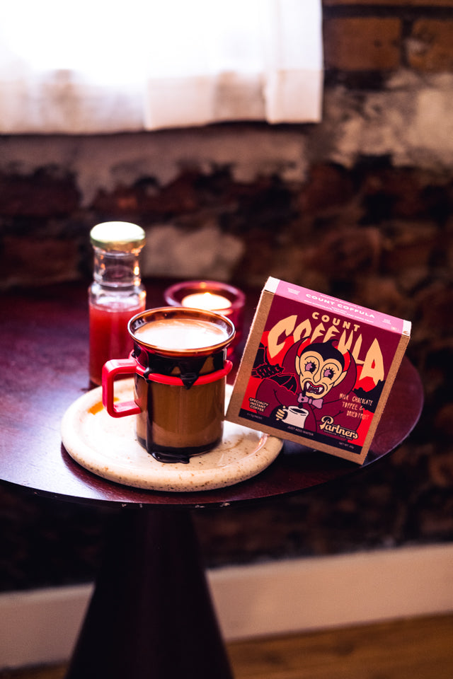 Halloween strawberry chocolate latte in a brown mug beside a Count Coffula instant coffee box and a syrup bottle on a small table with a candle and exposed brick wall