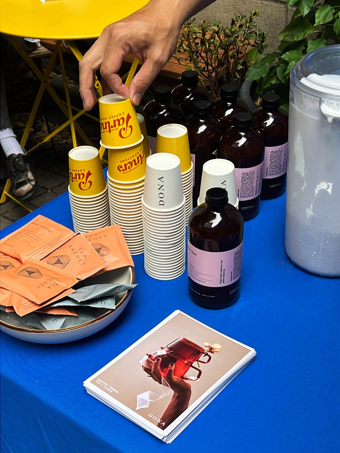 Hand picking up a yellow Dona cup beside stacked cups, bottled drinks, tea sachets, and a magazine on a blue table
