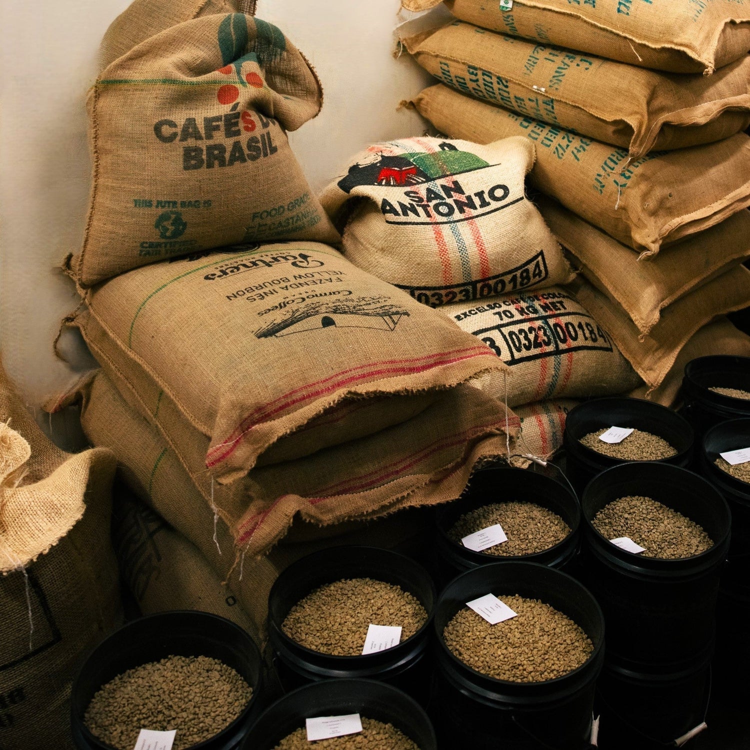 Partners Coffee image for A Note On Tariffs blog post. This image depicts bags of un-roasted green coffee beans in buckets ready for roasting alongside burlap bags filled with the same green coffee beans