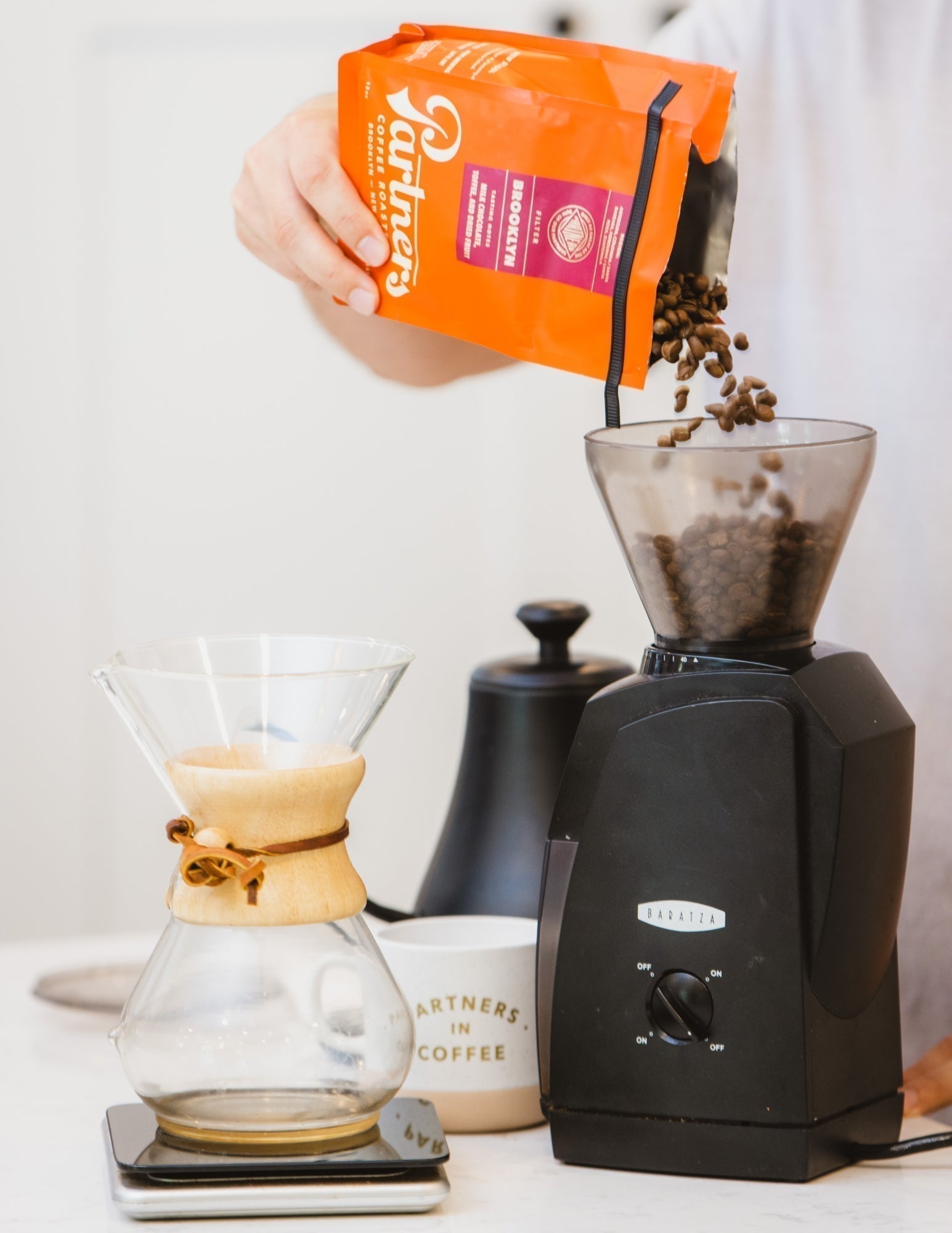 Partners Coffee Our Favorite Coffee Grinders blog post. This image depicts Partners Coffee's Brooklyn whole beans being poured into a Baratza grinder for use on a Chemex pour over.
