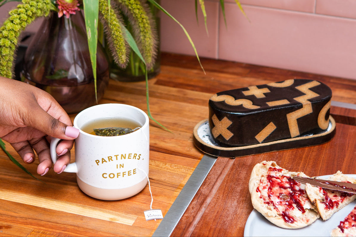 Hand holding a cup of tea with a tea sachet, cake, and jam toast on a wooden table