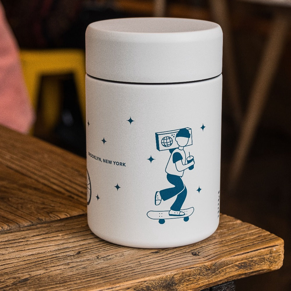 Three-quarter view of Party People MiiR coffee canister showing teal skateboarder illustration, twist bezel lid, and white hardshell powder coat on a wooden table