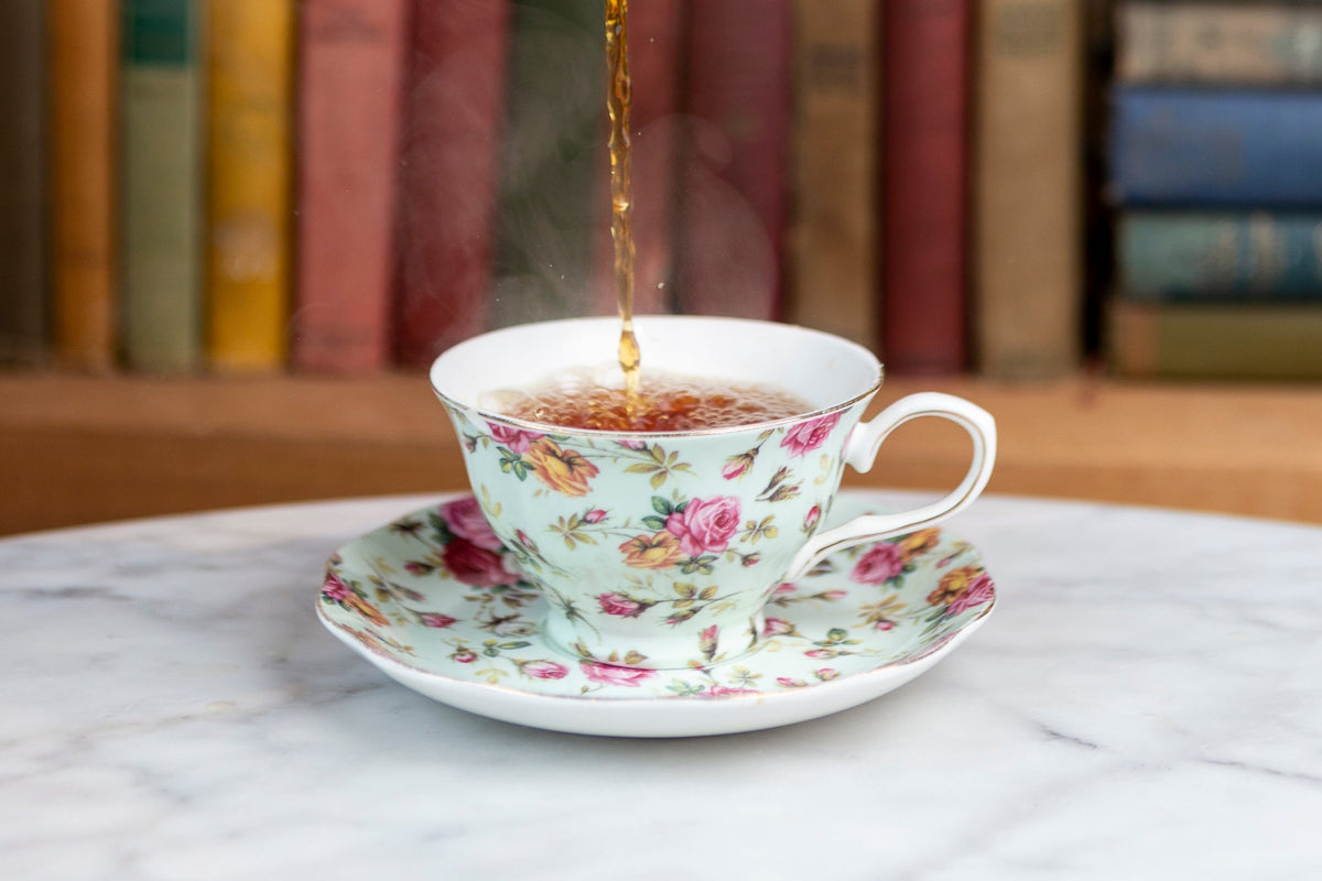 Tea being poured into a floral teacup on a saucer beside books