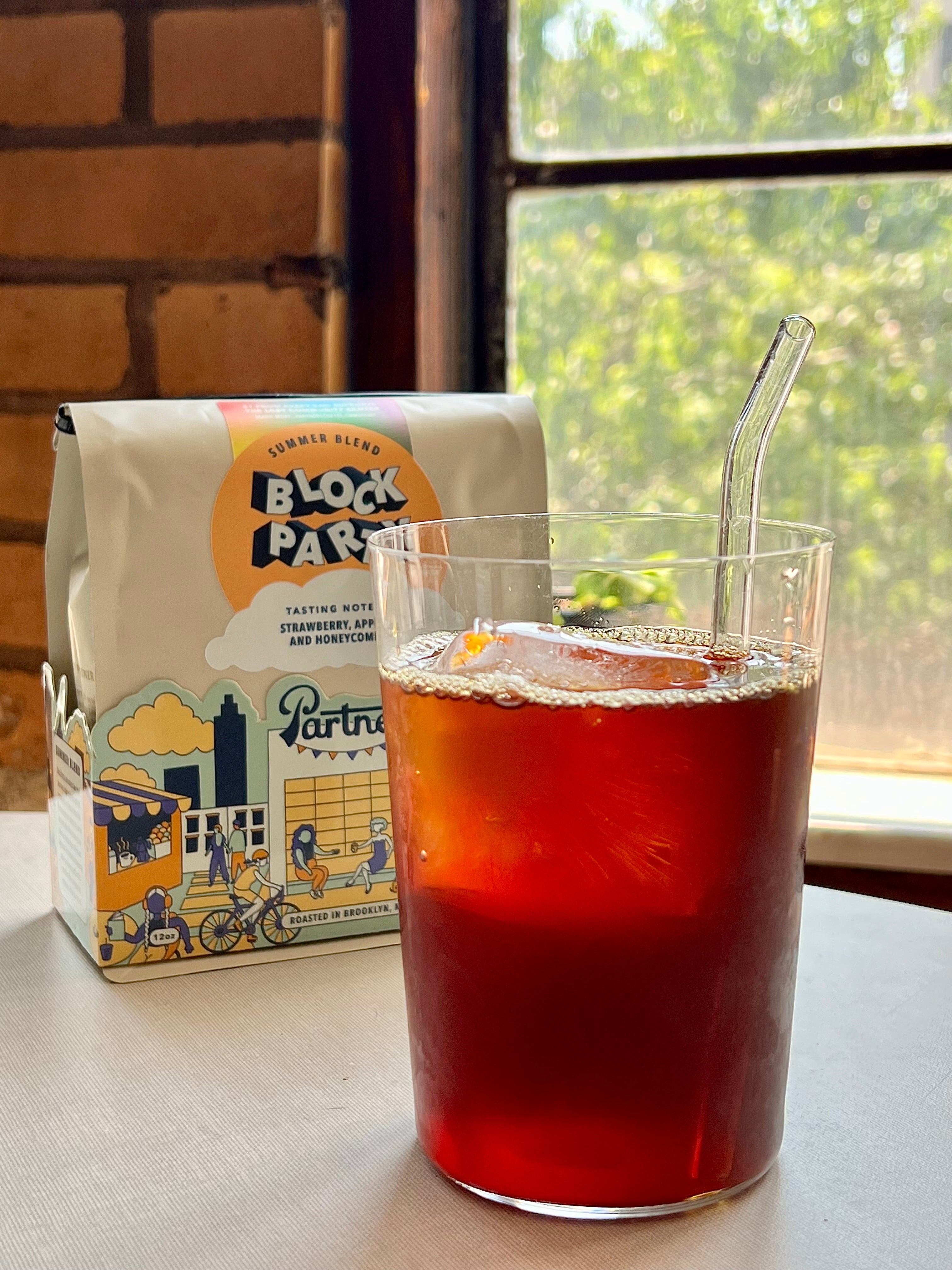 Image showing a cup of amber colored iced coffee in a glass with a straw, sitting in front of a white package of coffee with a blue and orange label reading "block party" with an illustration of new york city