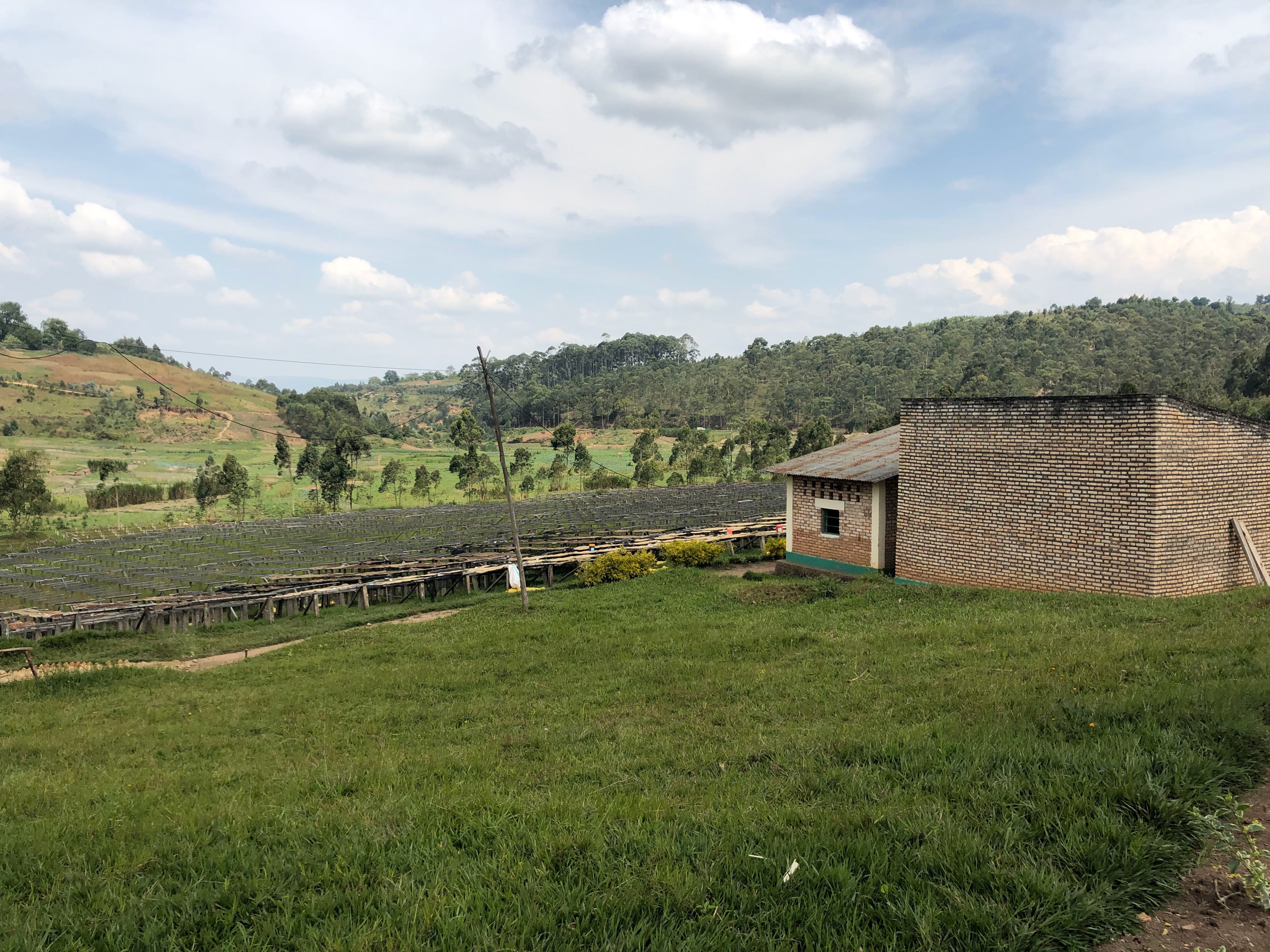Coffee drying beds and a brick processing building on a grassy hillside at Gahahe farm in Burundi under a partly cloudy sky