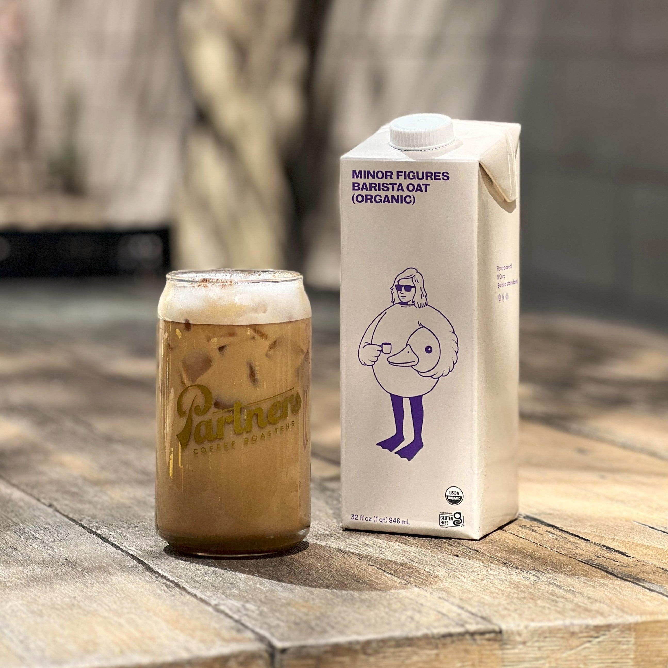 Iced Oatchata latte in a Partners Coffee glass beside a Minor Figures Barista Oat (organic) carton on a wooden table