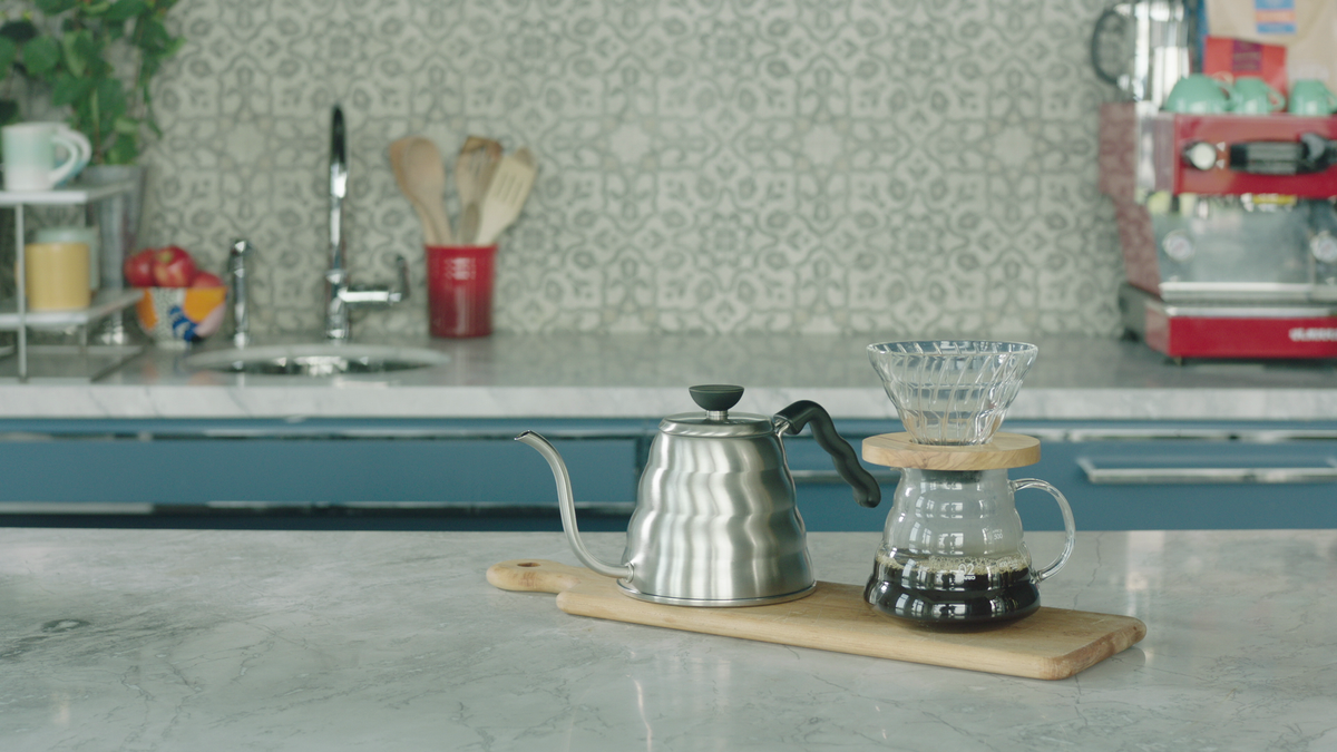 Stainless gooseneck kettle and Hario V60 dripper on a wooden board on a marble kitchen counter with tiled backsplash