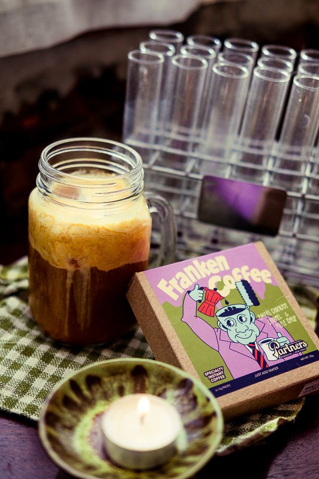 Mason jar of iced pumpkin coffee topped with pumpkin foam beside FrankenCoffee instant coffee box and lit candle on checkered napkin