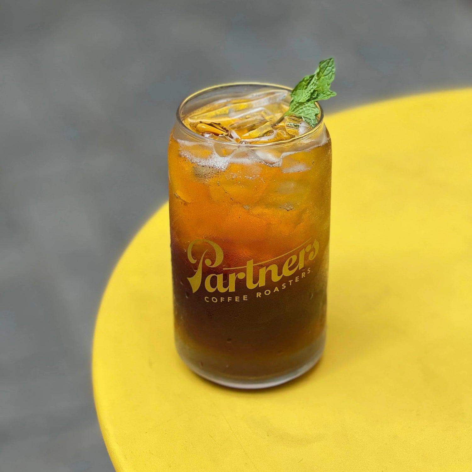 Iced vanilla cold brew soda in a Partners-branded glass with ice and a mint sprig on a yellow table