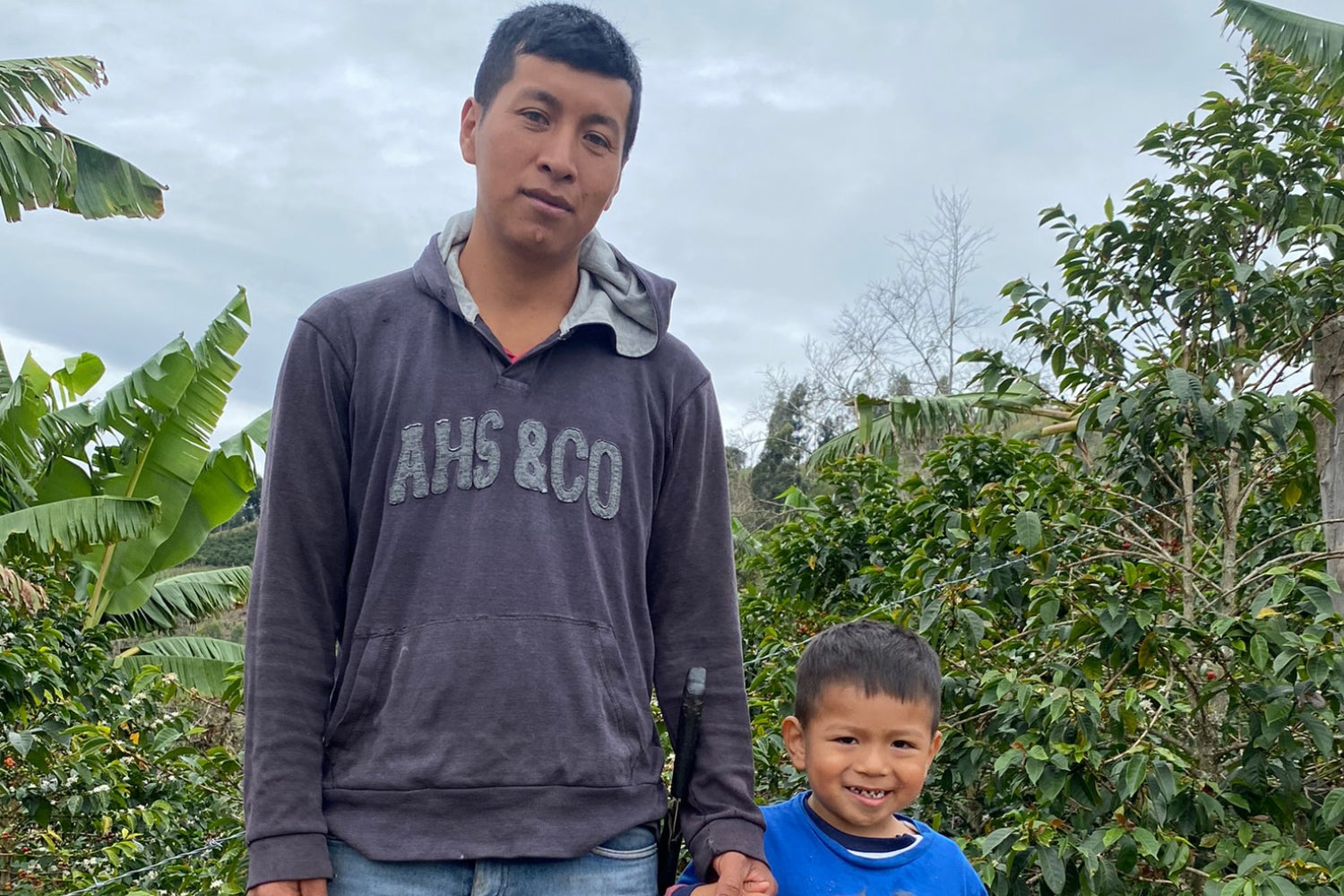 Vladimir Calambás standing with a young boy on his Colombian coffee farm with coffee plants and banana trees in the background