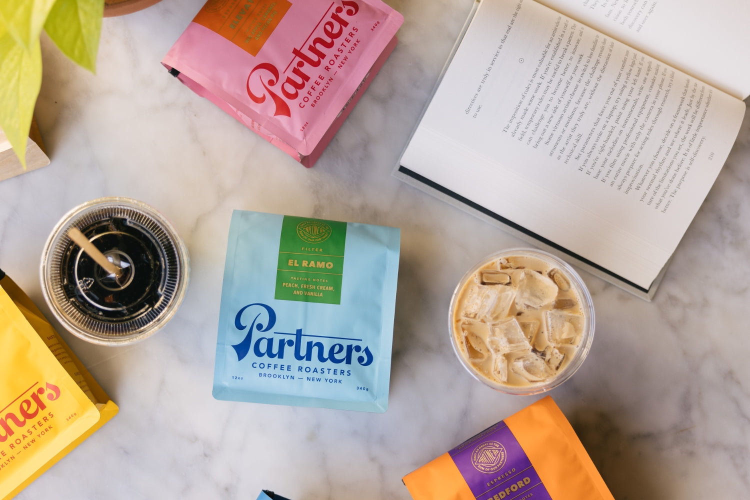 Overhead view of Partners Coffee bags with a cold brew, iced latte, and open book on a marble tabletop