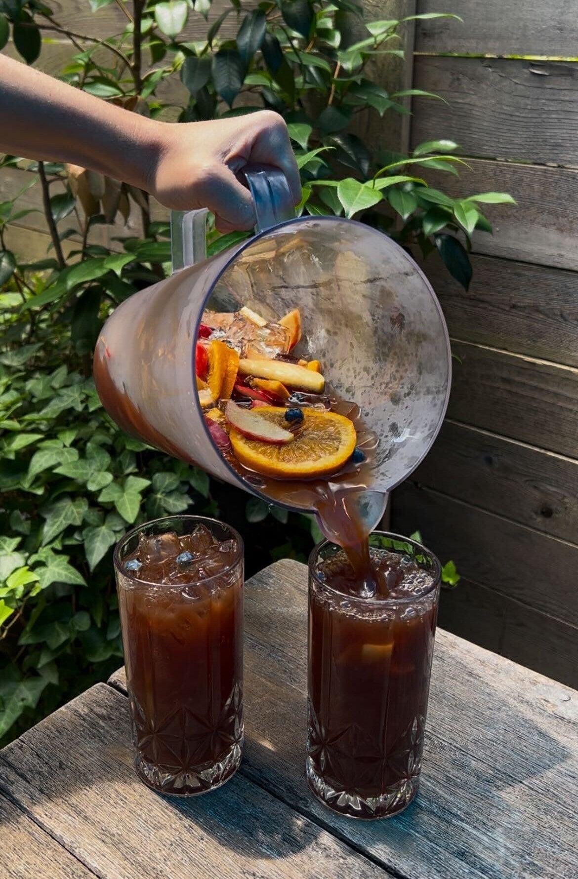 Hand pouring cold-brew sangria from a pitcher into two tall glasses on a weathered wooden table with citrus slices and berries visible