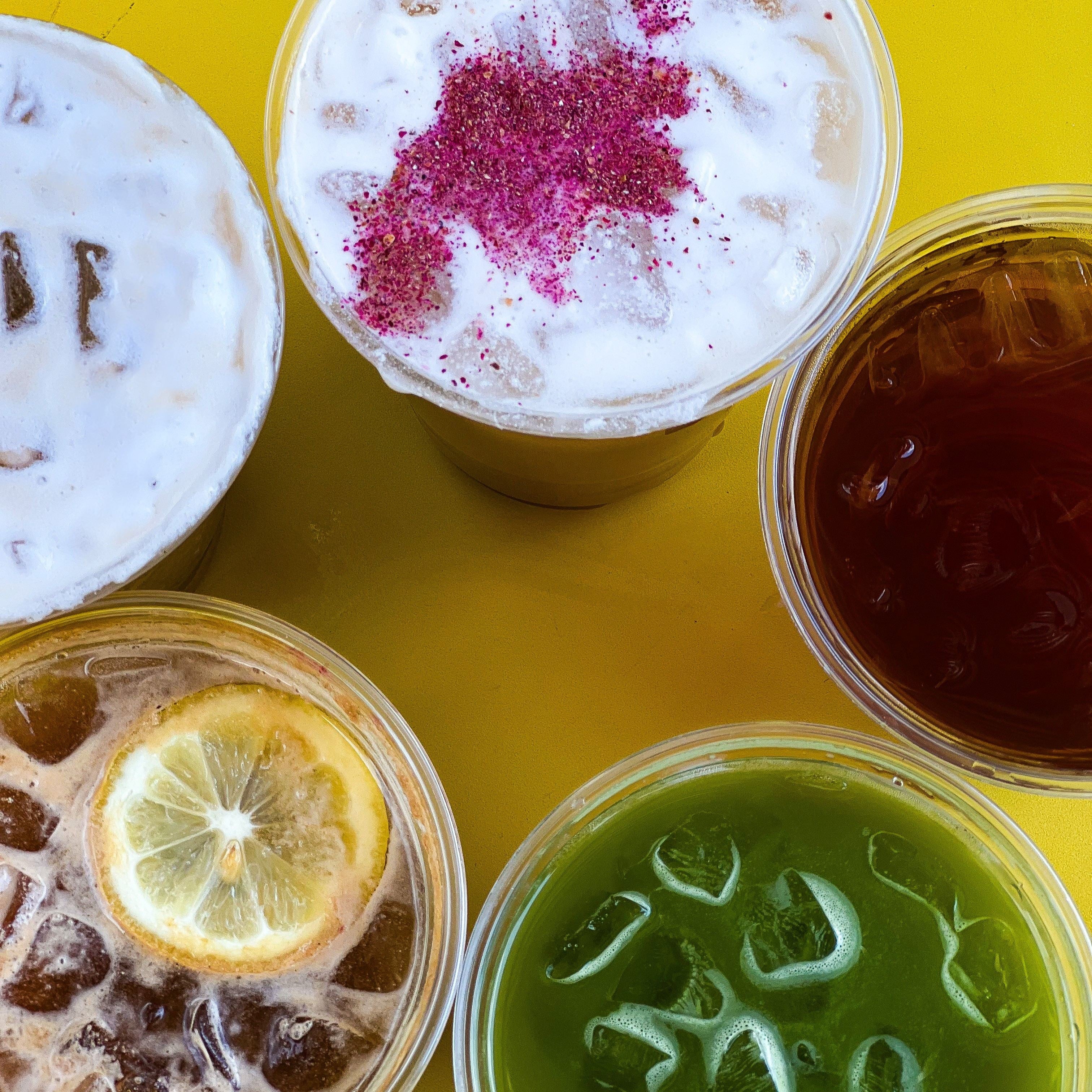 Five iced coffee drinks on a yellow table: lemon-topped drink, green matcha, dark iced coffee, and two foam-topped drinks