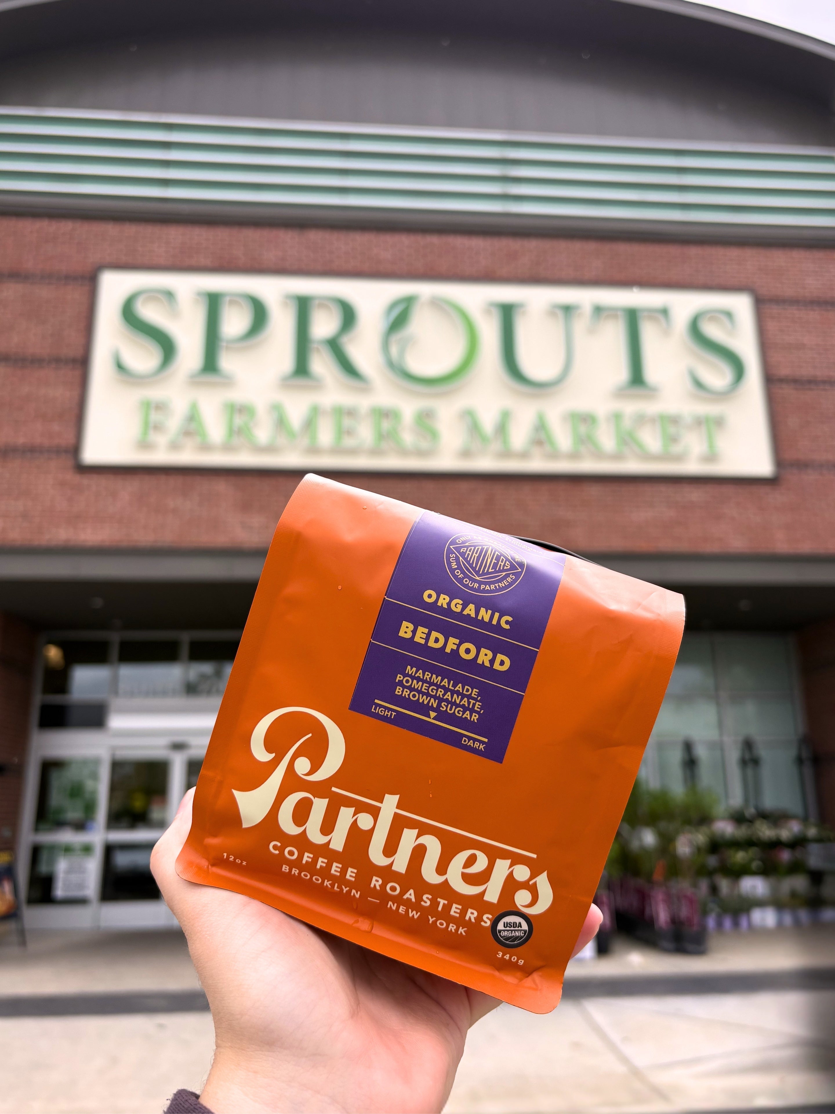 Hand holding an orange bag of Partners Coffee Organic Bedford in front of a Sprouts Farmers Market storefront