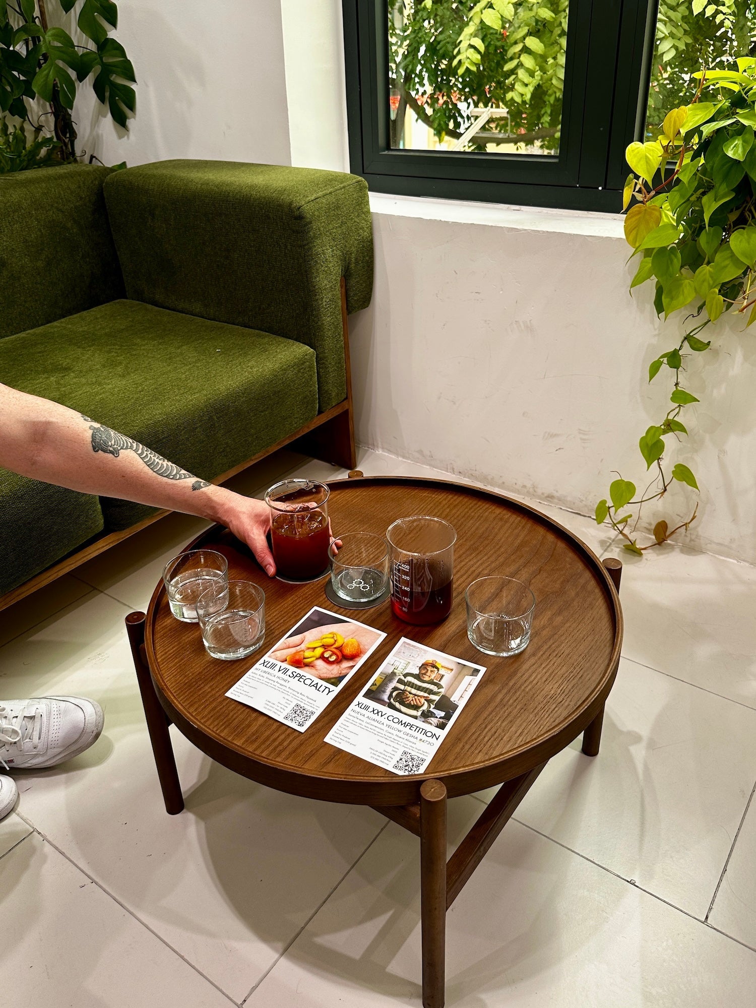 Two glass beakers of brewed coffee and tasting glasses on a round wooden table, hand reaching in, green sofa and window plants in a Hanoi café