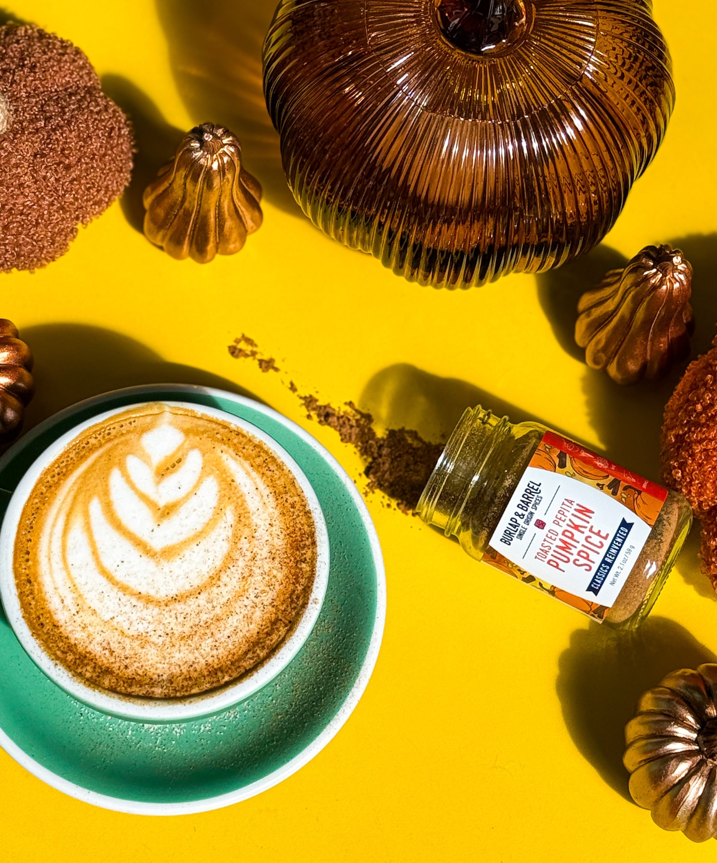 Burlap & Barrel x Partners Coffee recipe image. This image depicts the Pumpkin Pie Latte aside Burlap & Barrel's Toated Pepita Pumpkin Spice surrounded by seasonal themed  decorations for fall